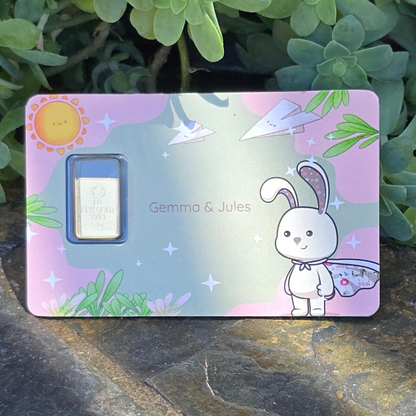 Gold bar in a decorative case with cartoon character and 'Gemma & Jules' branding, surrounded by green plants.